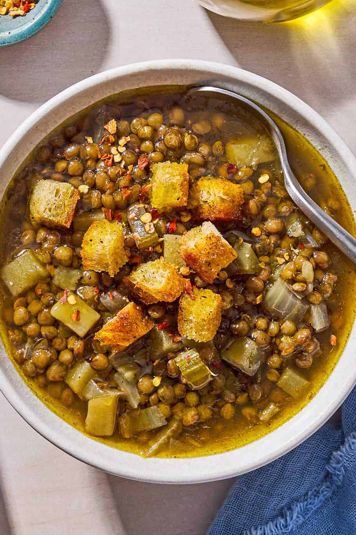 An overhead photo of a bowl of lentil potato soup with a spoon, topped with croutons. Next to this is a spoon, a cloth napkin, a bottle of olive oil, and a pot with the rest of the soup.