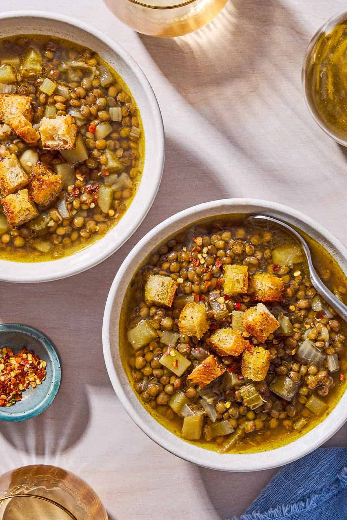 An overhead photo of 2 bowl of lentil potato soup topped with croutons, one with a spoon. Next to these are 2 glasses of water, a bottle of olive oil, and a bowl of red pepper flakes.