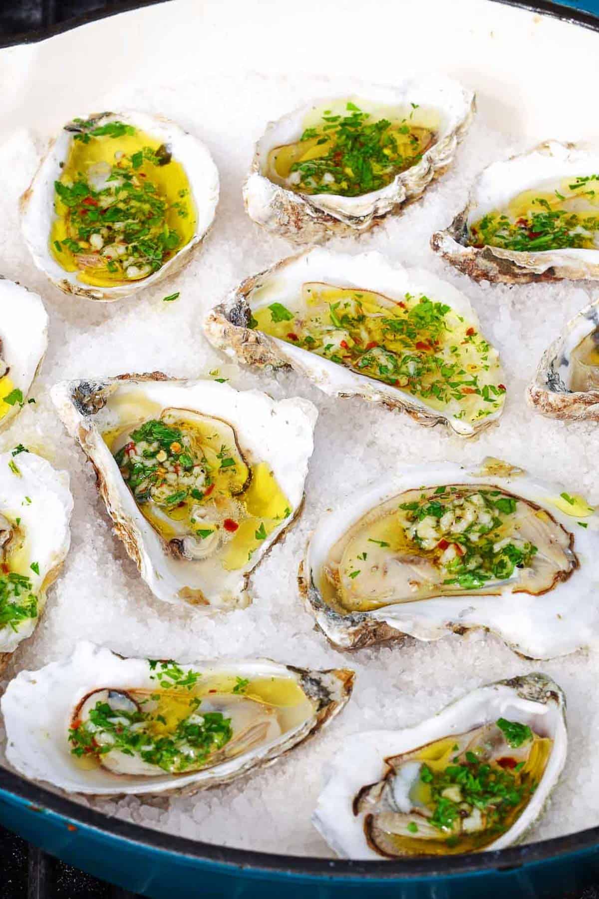 A close up of several grilled oysters with garlicky herb sauce on a bed of rock salt in a skillet.