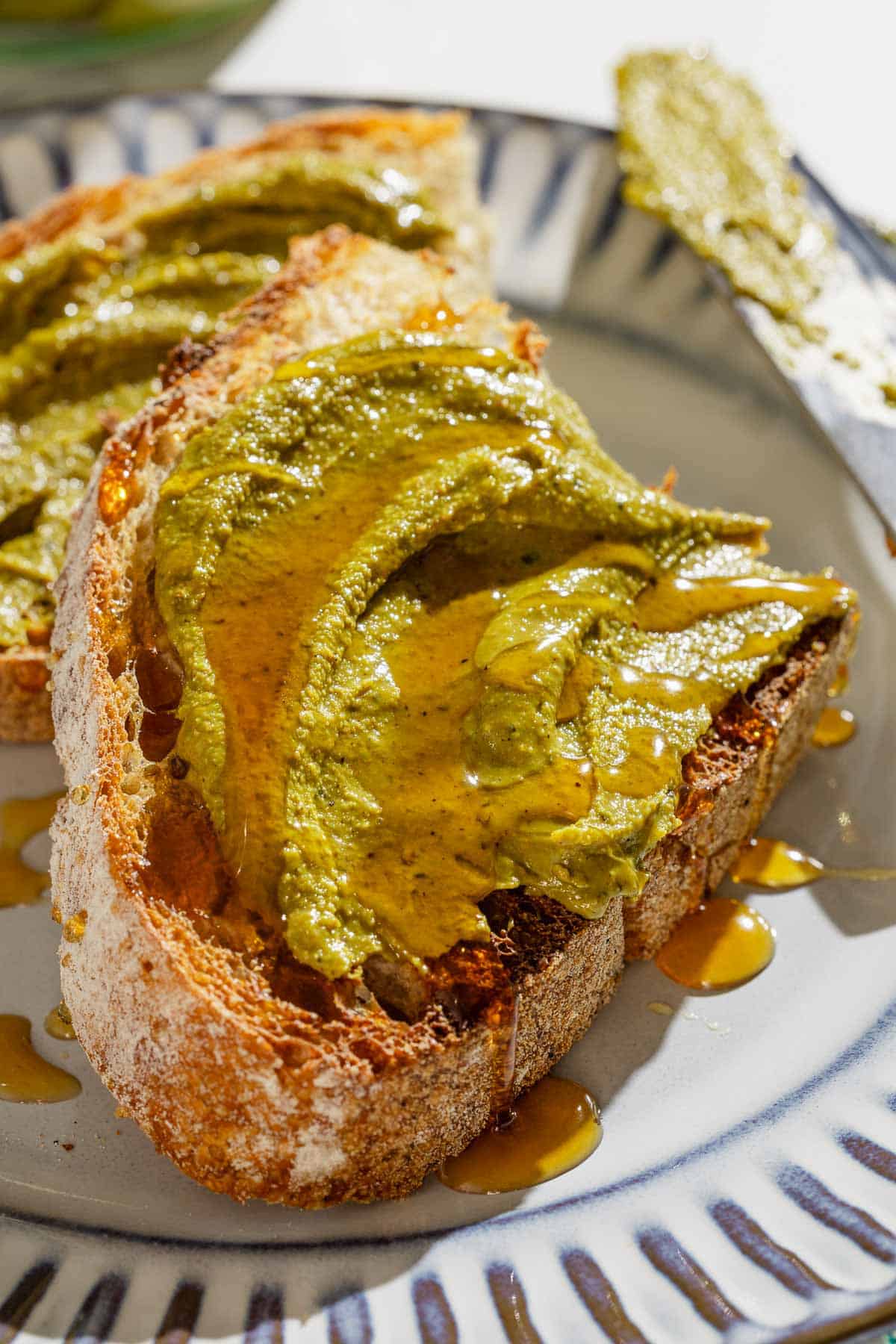 A close up of 2 halves of toast with pistachio butter drizzled with honey on a plate with a knife.