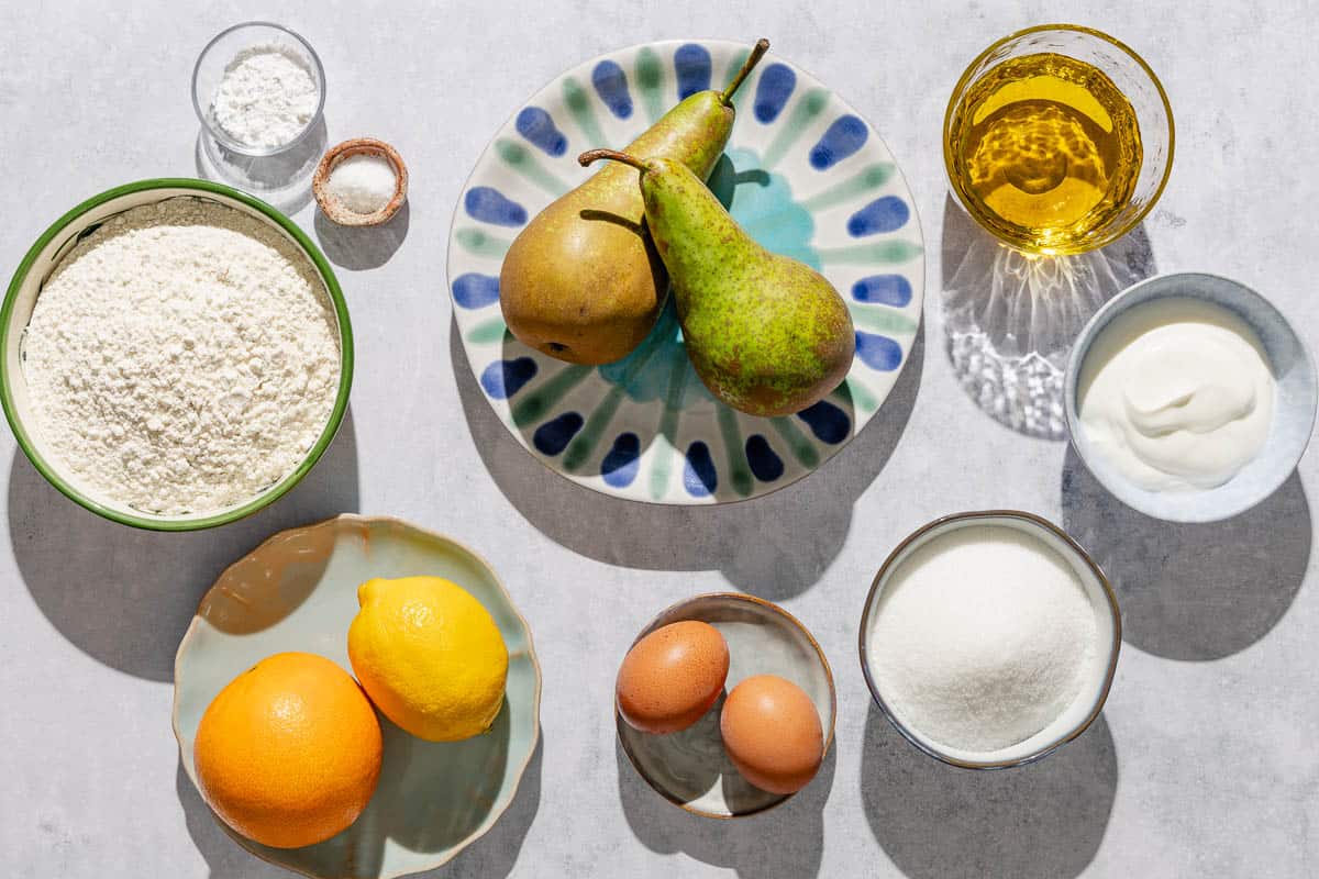 Ingredients for Italian pear cake including pears, flour, baking powder, salt, eggs, sugar, lemon, orange, olive oil, greek yogurt, vanilla extract and powdered sugar.