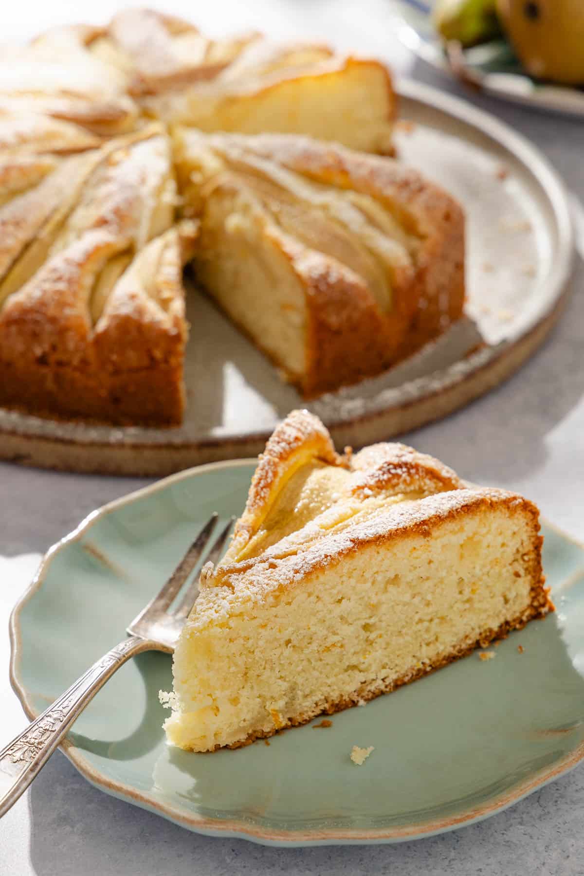 a slice of Italian pear cake on a plate with a fork, with the rest of the cake on a platter in the background.