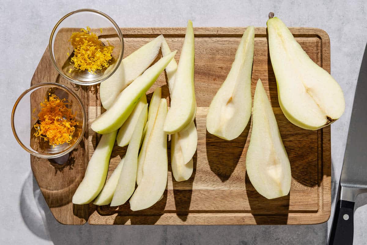 An overhead photo of sliced pears on a cutting board with a small bowl of lemon zest and a small bowl of orange zest. Next to this is a knife.