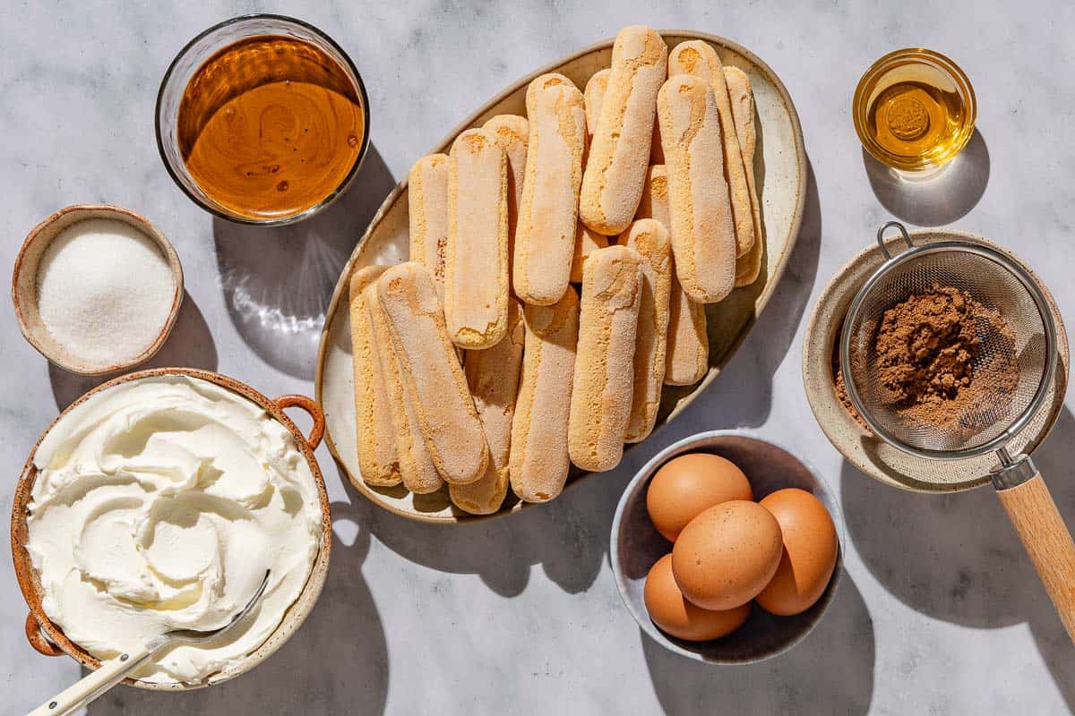 ingredients for tiramisu including eggs, sugar, mascarpone cheese, espresso, dark rum, ladyfinger cookies, and cocoa powder.