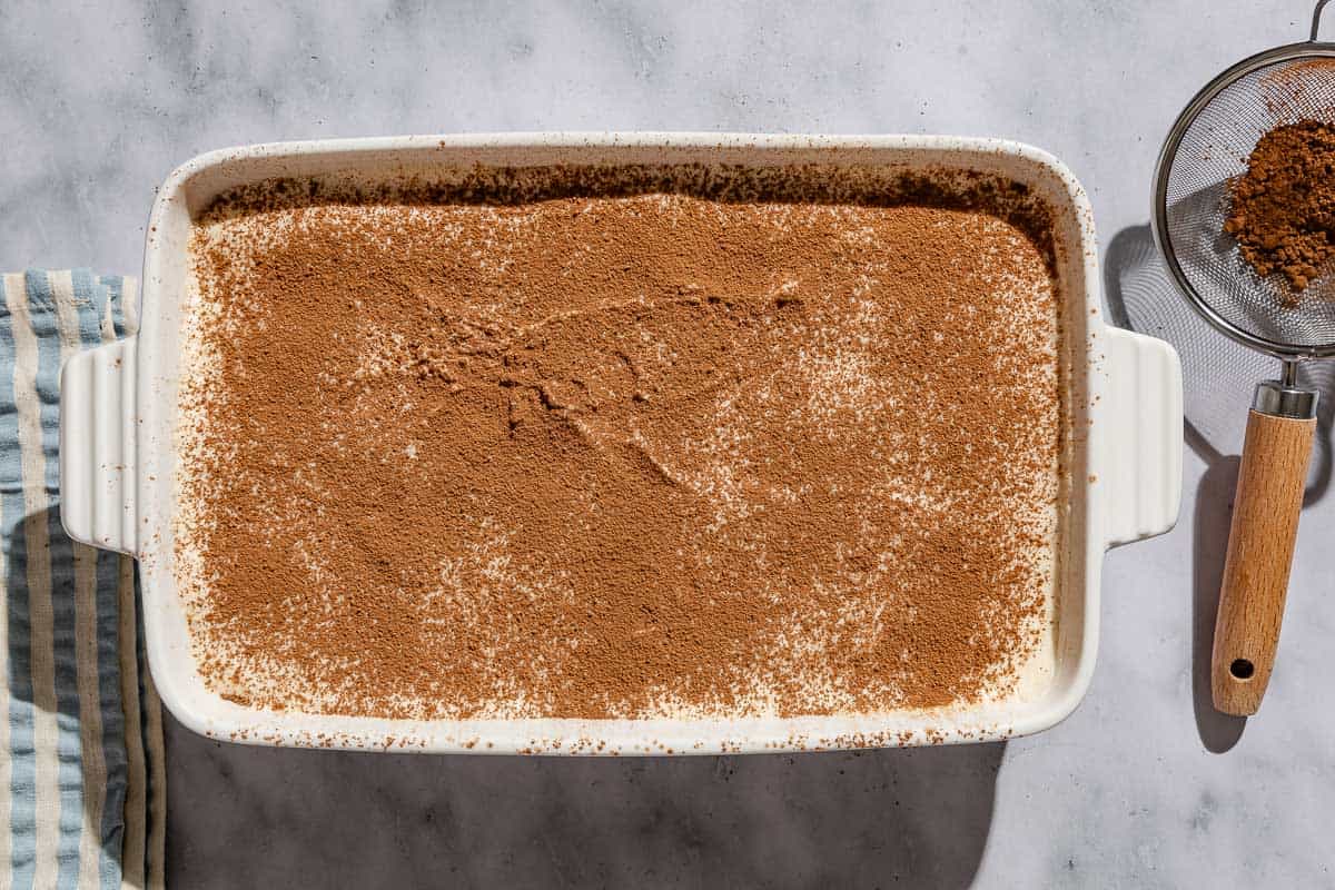 An overhead photo of the tiramisu in a baking dish just after being dusted with cocoa powder. Next to this is a sifter with the the cocoa powder.