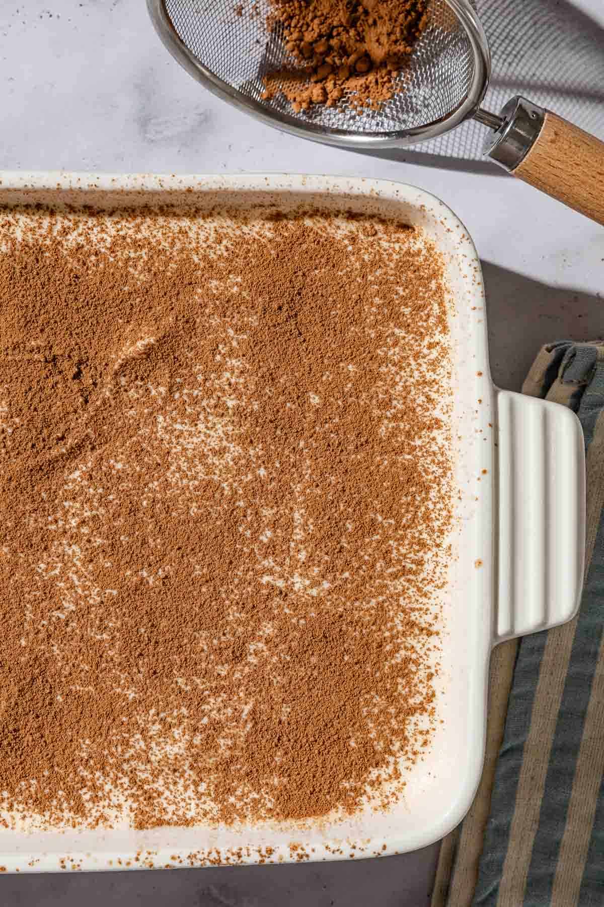 An overhead close up photo of the tiramisu in a baking dish just after being dusted with cocoa powder. Next to this is a sifter with the the cocoa powder.