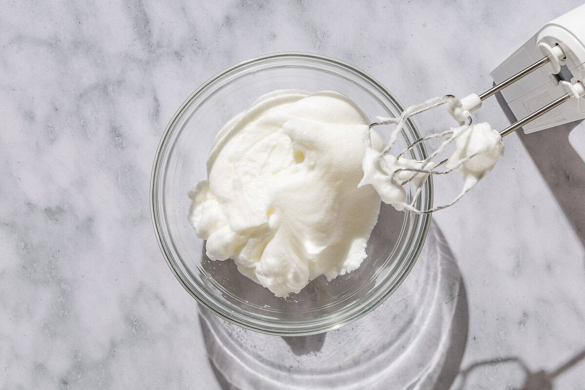 Egg whites beaten into stiff peaks in a bowl. Next to this is a hand mixer