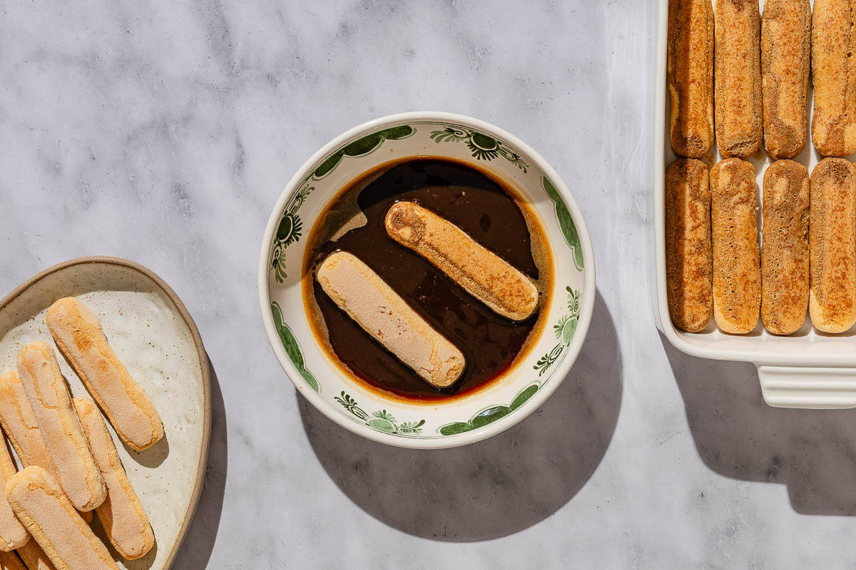 An overhead photo of several ladyfingers on a plate, 2 ladyfingers in a bowl of the espresso, and a baking dish with one layer of the ladyfingers evenly spread across the bottom.