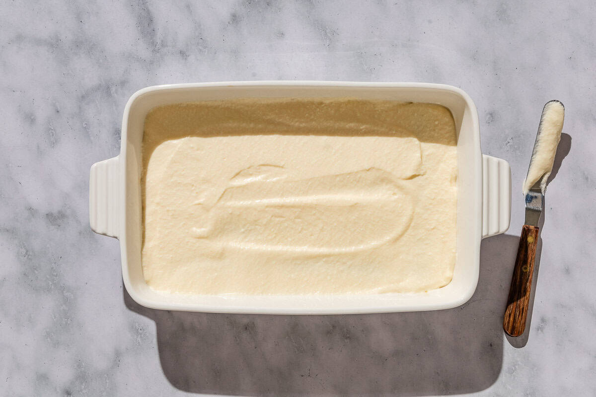 An overhead photo of the mascarpone mixture covering the first layer of lady fingers in a baking dish. Next to this is a knife.