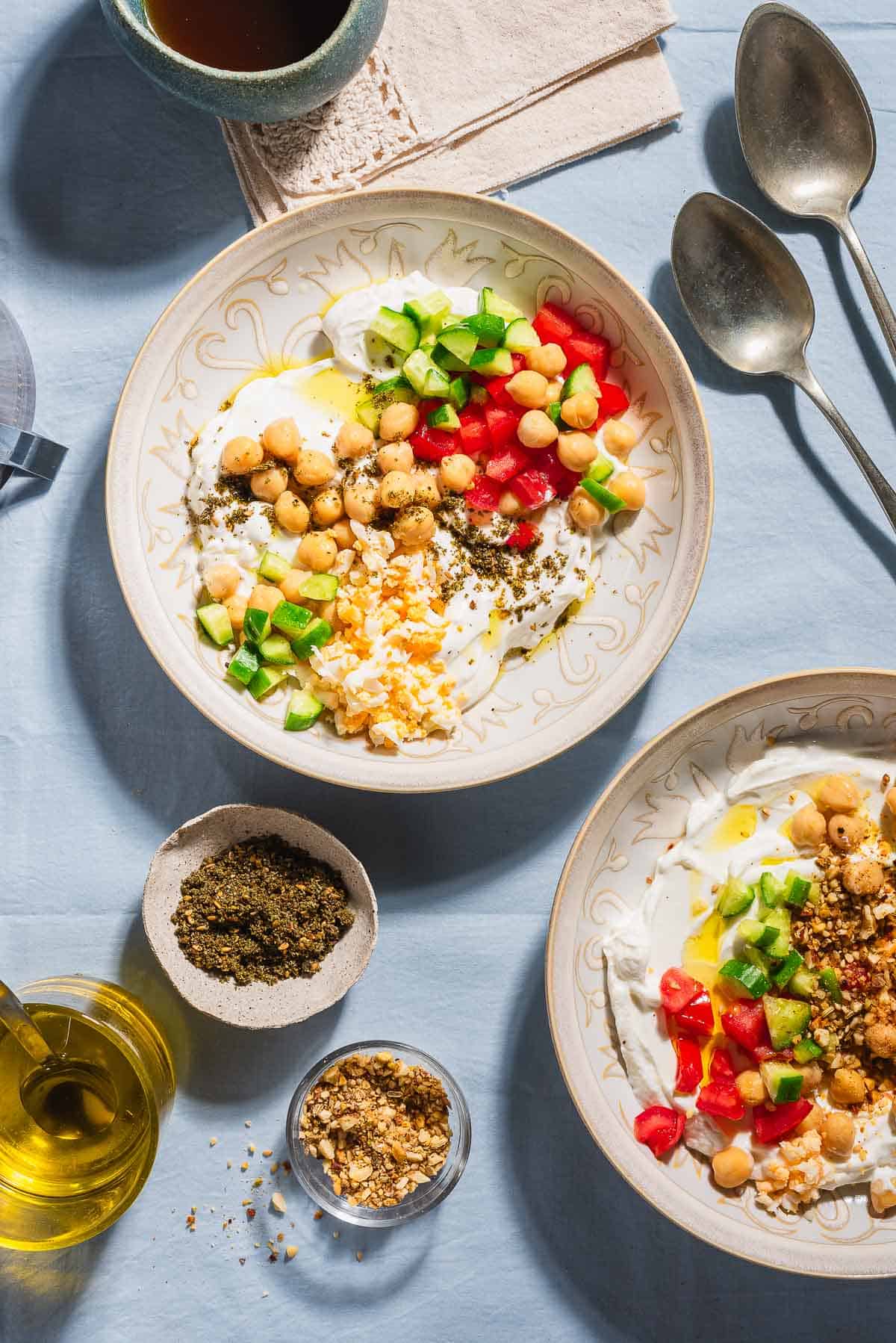 An overhead photo of 2 savory yogurt bowls. Next to these are bowls of dukkah and za'atar, 2 spoons, a cup of coffee, and a cup of olive oil with a spoon.