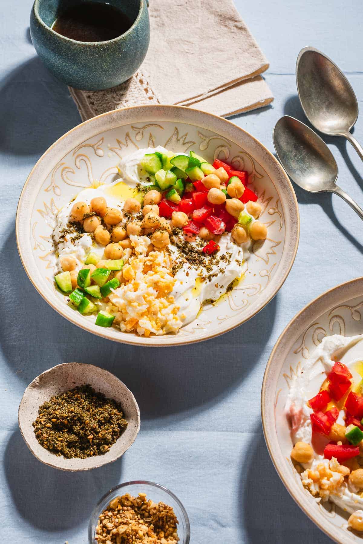 A close up of 2 savory yogurt bowls. Next to these are bowls of dukkah and za'atar, 2 spoons, and a cup of coffee.