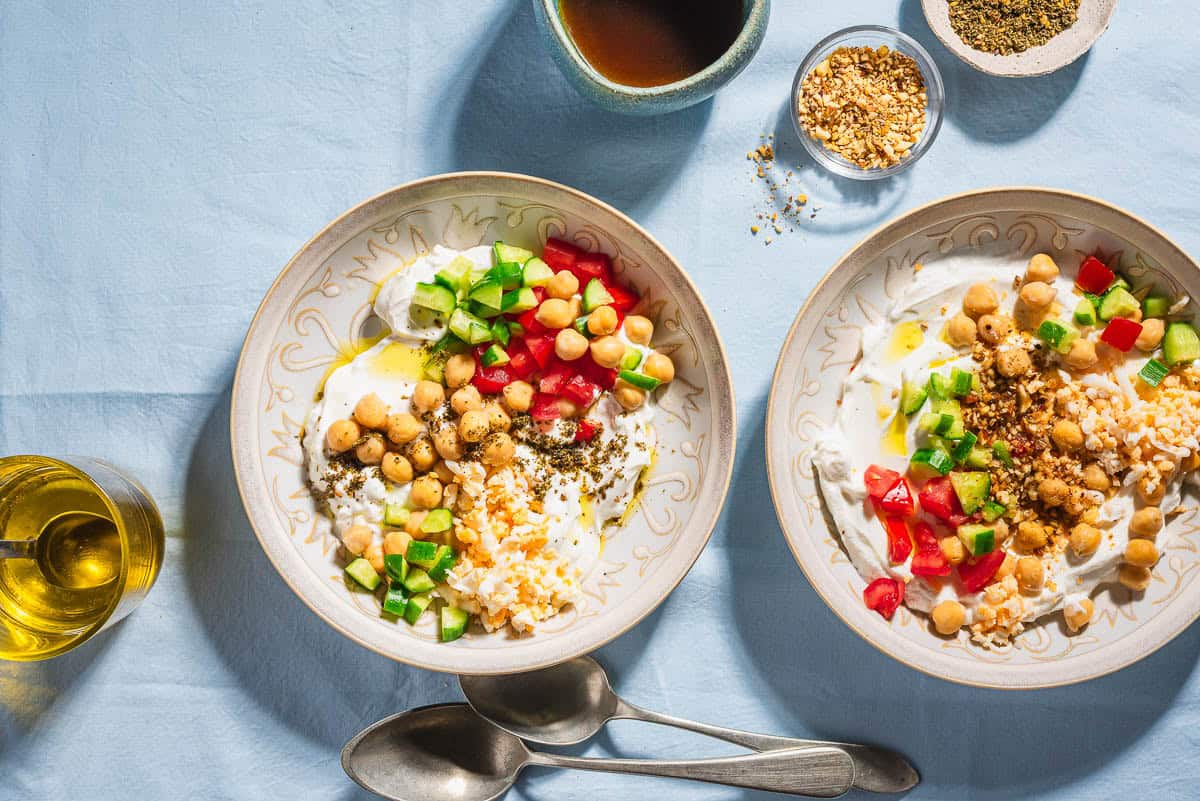 A close up of 2 savory yogurt bowls. Next to these are bowls of dukkah and za'atar, 2 spoons, a cup of coffee, and a cup of olive oil with a spoon.