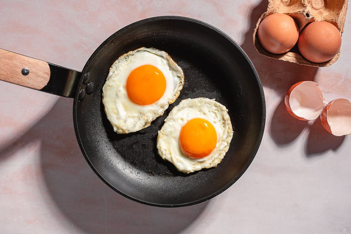 An overhead photo of 2 fried eggs in a skillet next to 2 eggs in a carton, and 2 eggshell halves.