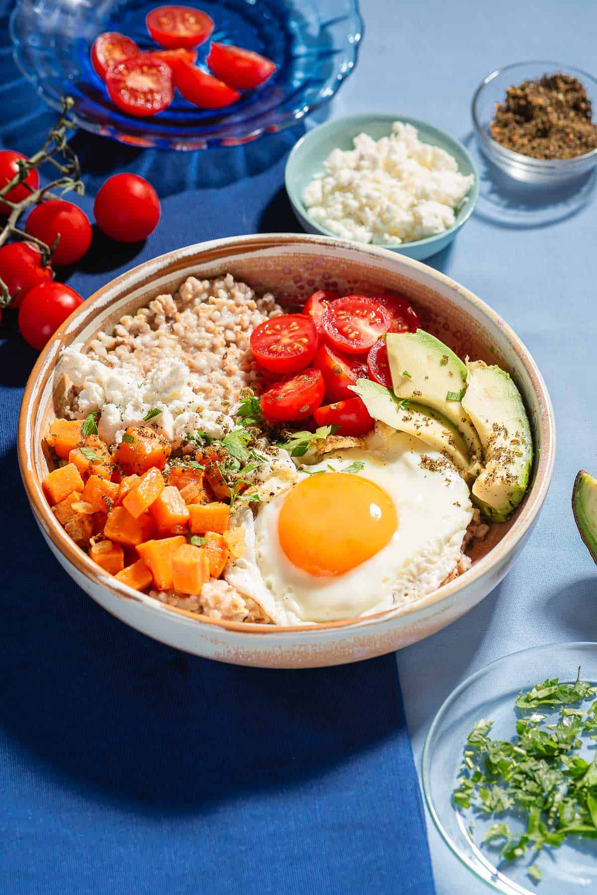 A savory oatmeal bowl surrounded by small bowls of feta and za'atar, cherry tomatoes, and chopped parsley.