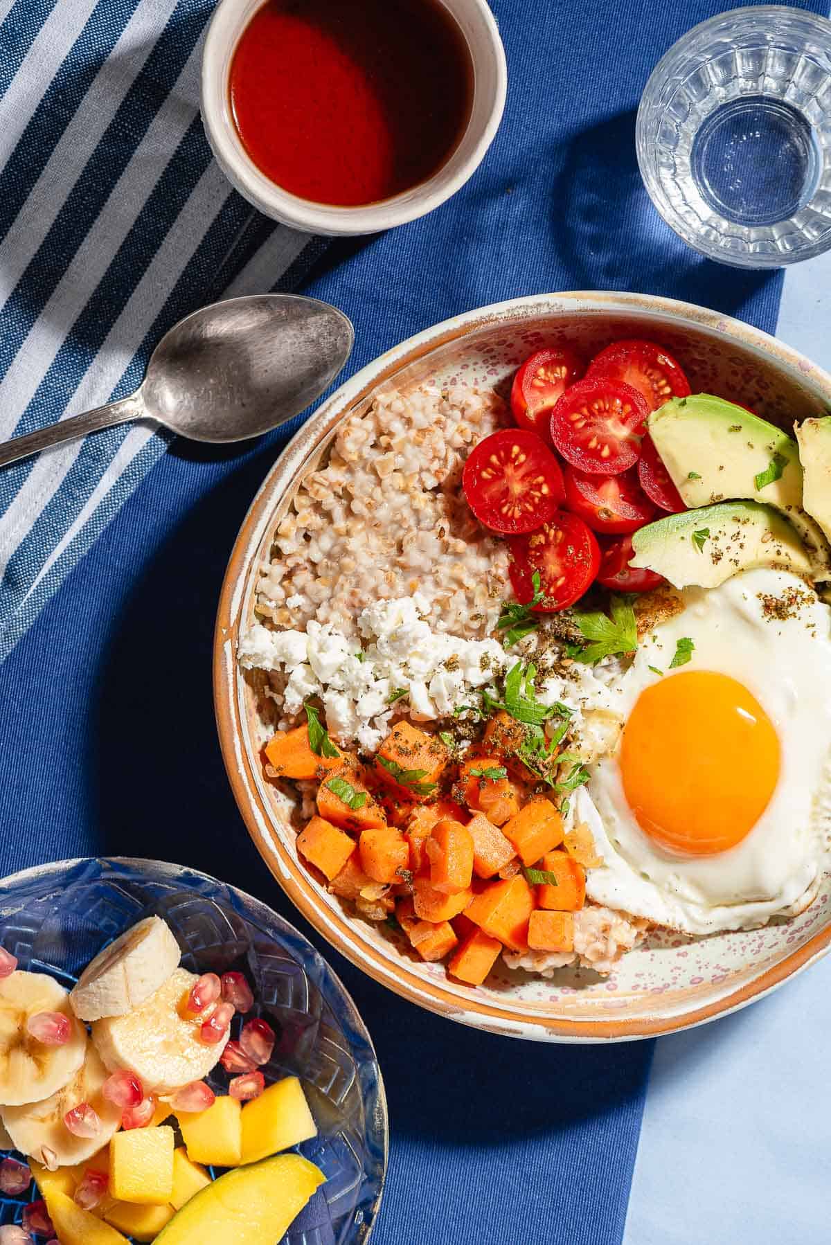 An overhead photo of a savory oatmeal bowl surrounded by a bowl of fruit, a spoon, a glass of water and a cup of tea.