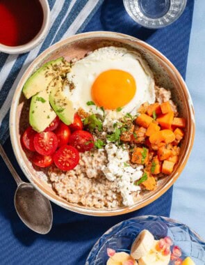 An overhead photo of a savory oatmeal bowl surrounded by a bowl of fruit, a spoon, a glass of water and a cup of tea.