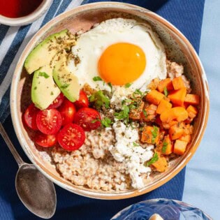 An overhead photo of a savory oatmeal bowl surrounded by a bowl of fruit, a spoon, a glass of water and a cup of tea.