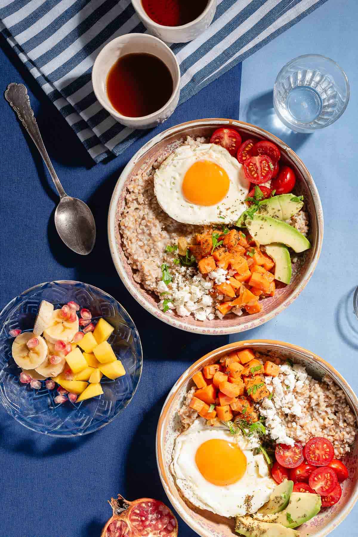 An overhead photo of a 2 savory oatmeal bowls surrounded by a bowl of fruit, a spoon, a glass of water and 2 cups of tea.