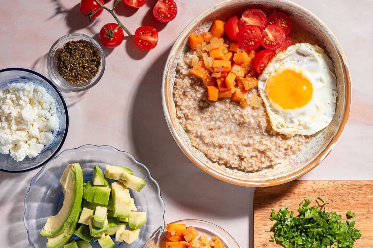 An overhead photo of a savory oatmeal bowl surrounded by small bowls of feta and za'atar, cherry tomatoes, chopped parsley, avocado and sauteed sweet potatoes and onions.