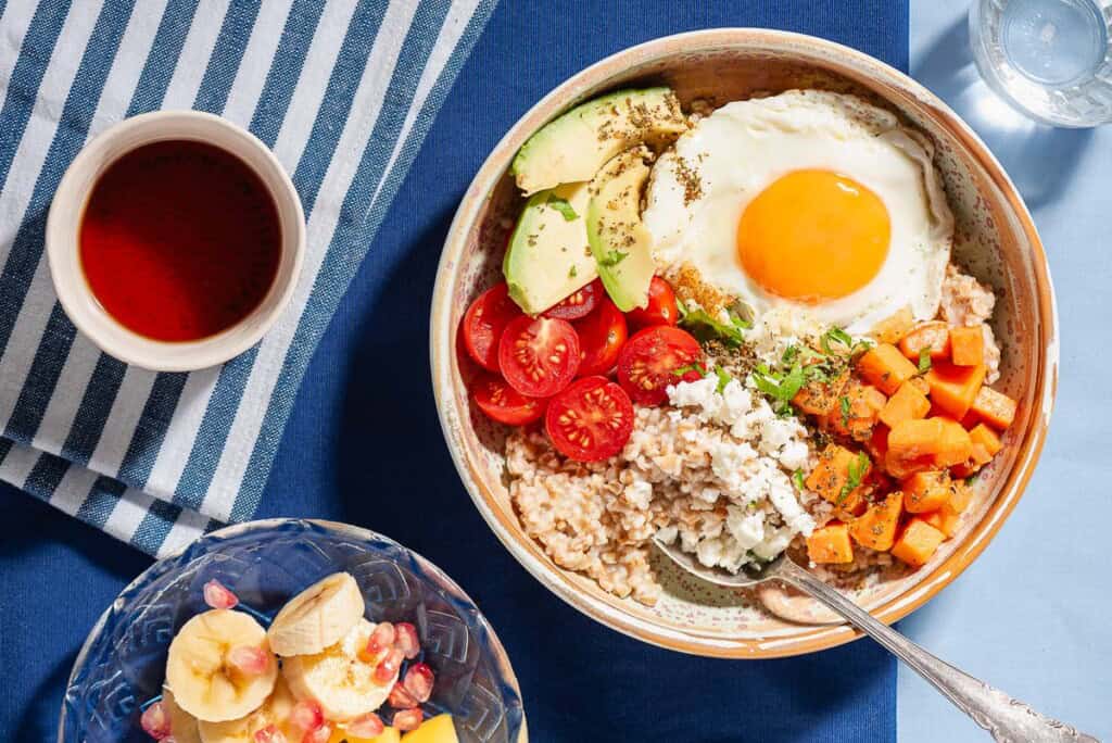 An overhead photo of a savory oatmeal bowl with a spoon surrounded by a bowl of fruit, a glass of water and a cup of tea.