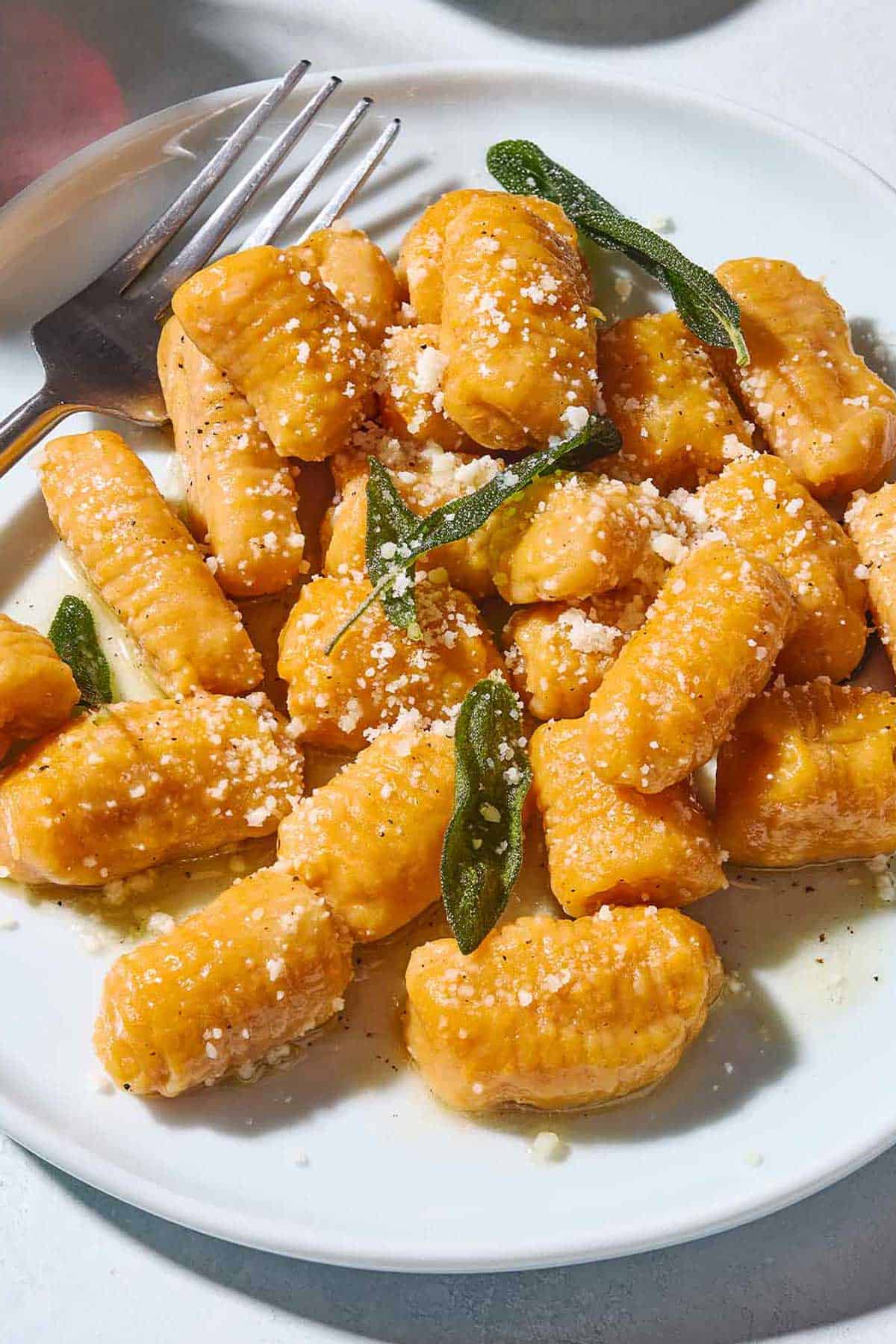 A close up of sweet potato gnocchi and sauce topped with fresh sage and parmigiano-reggiano cheese on a plate with a fork.