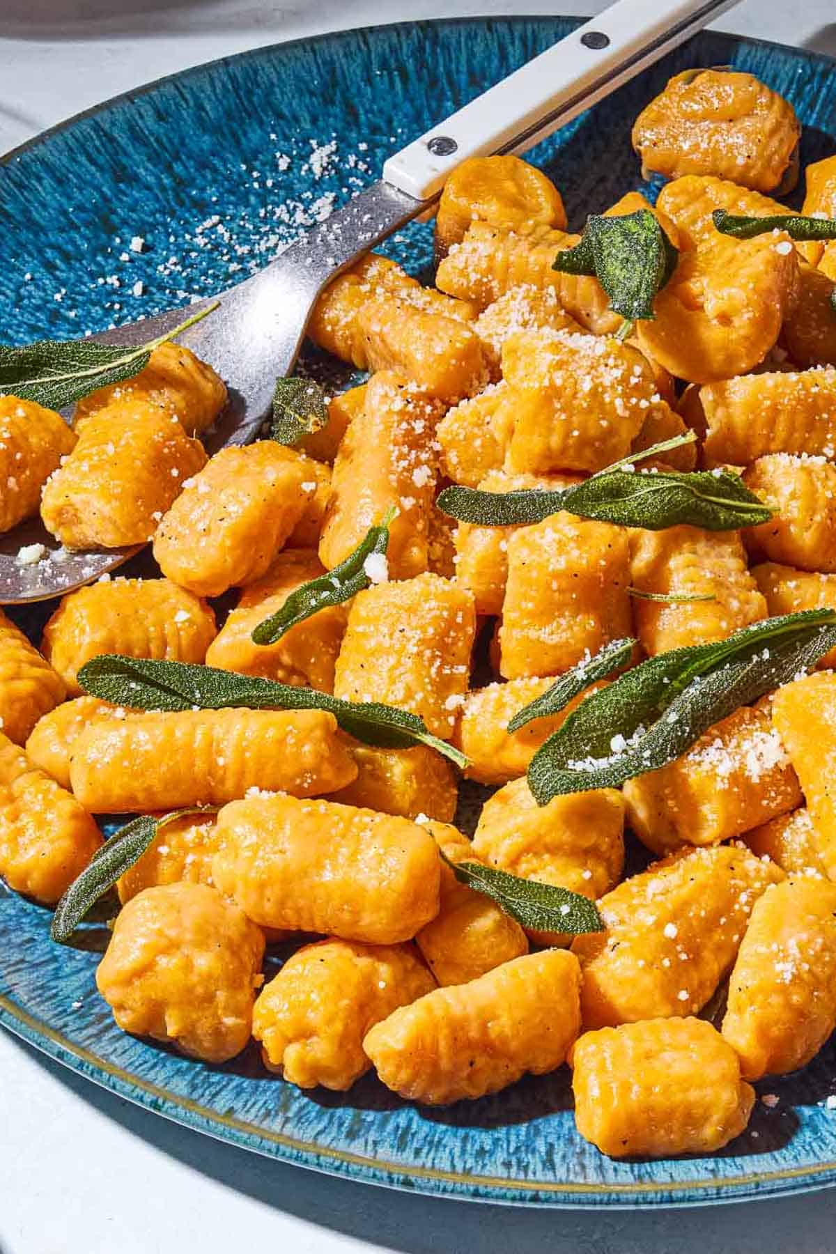 A close up of sweet potato gnocchi and sauce topped with fresh sage on a serving platter with a spoon.