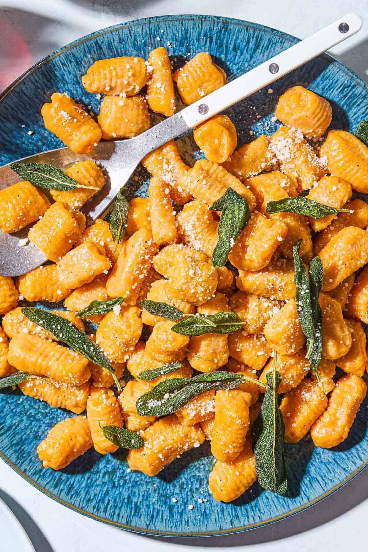 A close up of sweet potato gnocchi and sauce topped with fresh sage on a serving platter with a spoon.