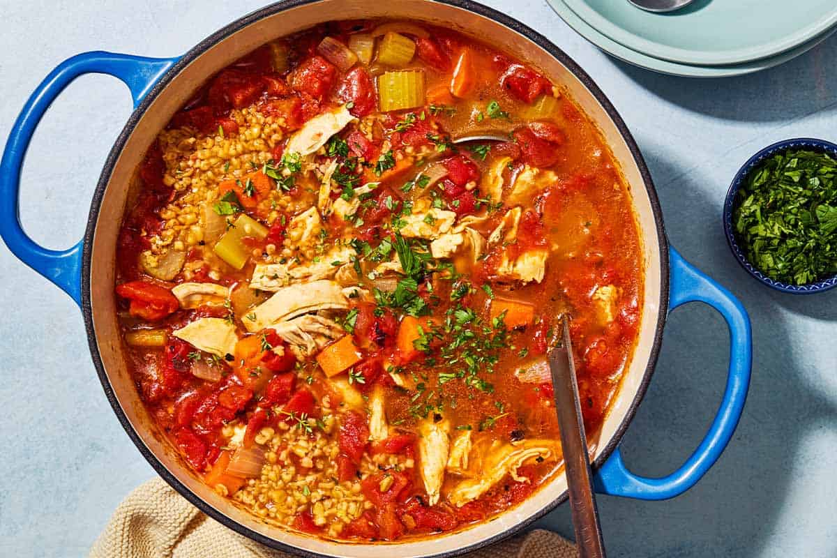 An overhead photo of freekeh soup with chicken in a large pot with a ladle. Next to this is a small bowl of chopped parsley.