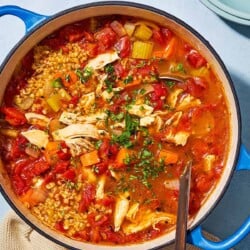 An overhead photo of freekeh soup with chicken in a large pot with a ladle. Next to this is a small bowl of chopped parsley, a stack of bowls with a spoon on top, and a kitchen towel.