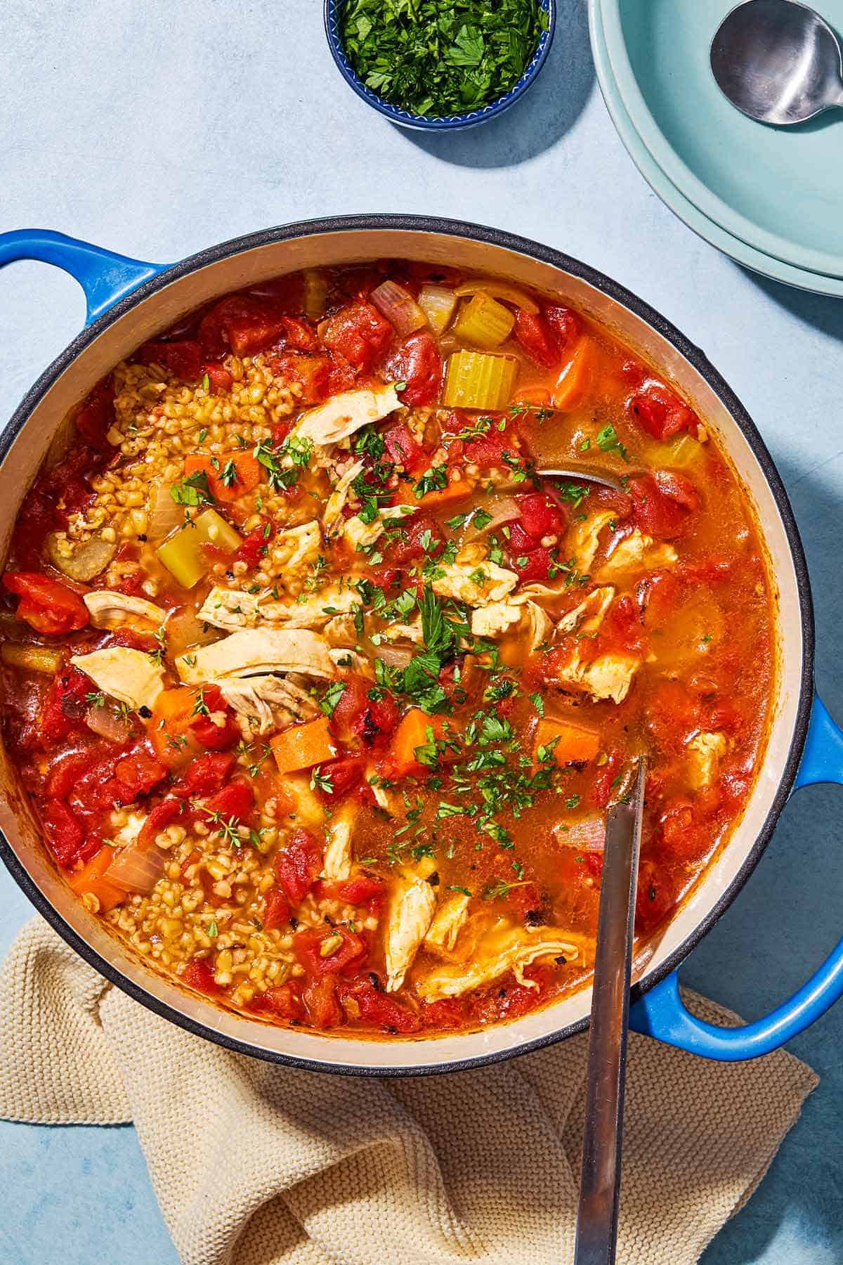 An overhead photo of freekeh soup with chicken in a large pot with a ladle. Next to this is a small bowl of chopped parsley, a stack of bowls with a spoon on top, and a kitchen towel.