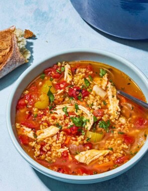 Freekeh soup with chicken in a bowl with a spoon. Next to this is a pot of the soup and some crusty bread.