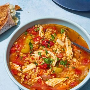 Freekeh soup with chicken in a bowl with a spoon. Next to this is a pot of the soup and some crusty bread.