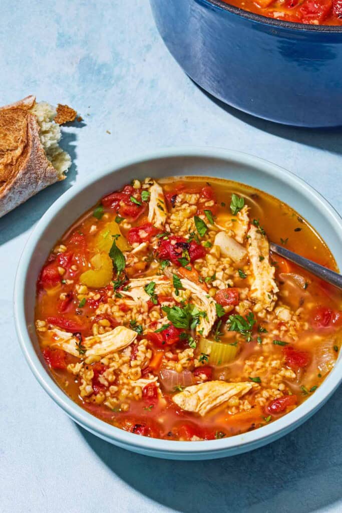 Freekeh soup with chicken in a bowl with a spoon. Next to this is a pot of the soup and some crusty bread.