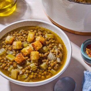 A bowl of lentil potato soup topped with croutons. Next to this is a spoon, a cloth napkin, a bottle of olive oil, and a pot with the rest of the soup.