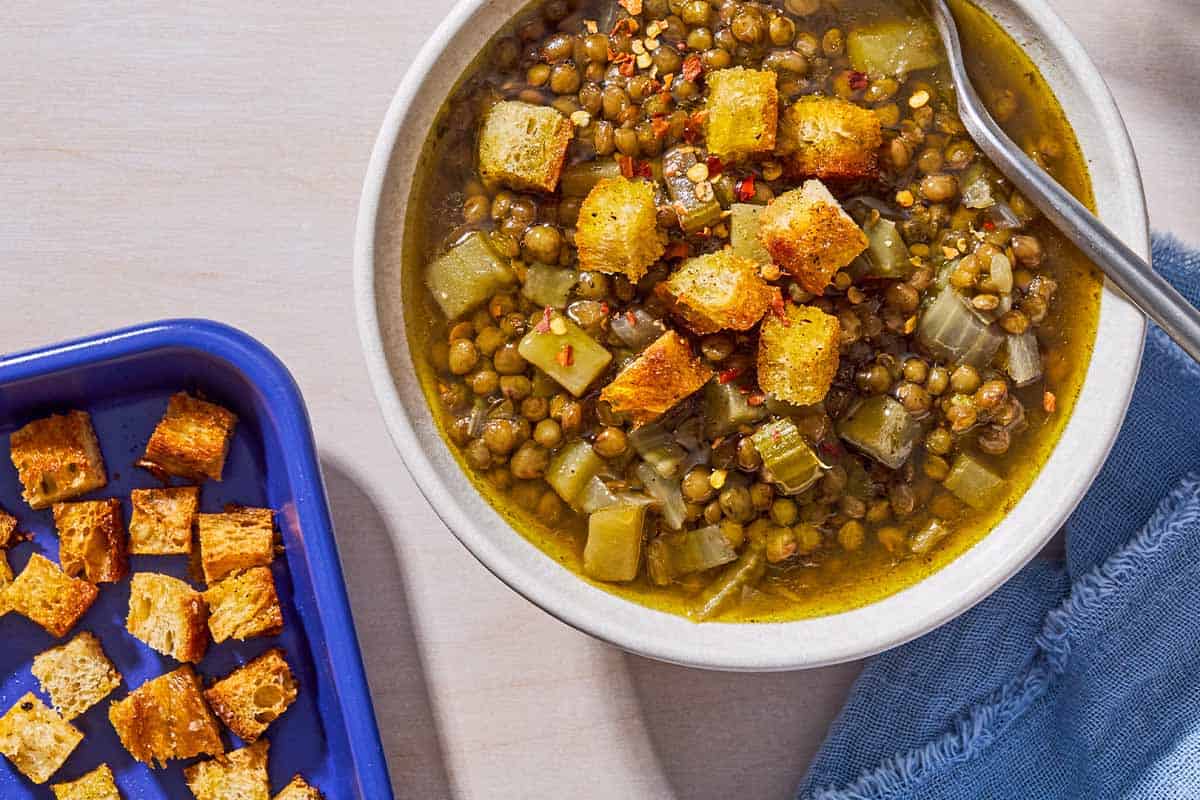 An overhead photo of a bowl of lentil potato soup with a spoon, topped with croutons. Is a cloth napkin and a baking sheet with the rest of the croutons.