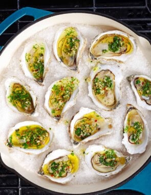 An overhead photo of several grilled oysters with garlicky herb sauce on a bed of rock salt in a skillet sitting on a grill.