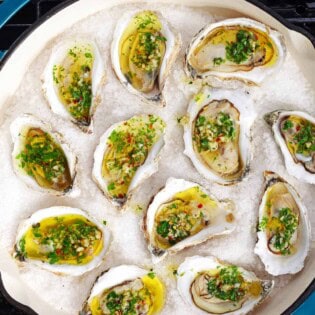 An overhead photo of several grilled oysters with garlicky herb sauce on a bed of rock salt in a skillet sitting on a grill.