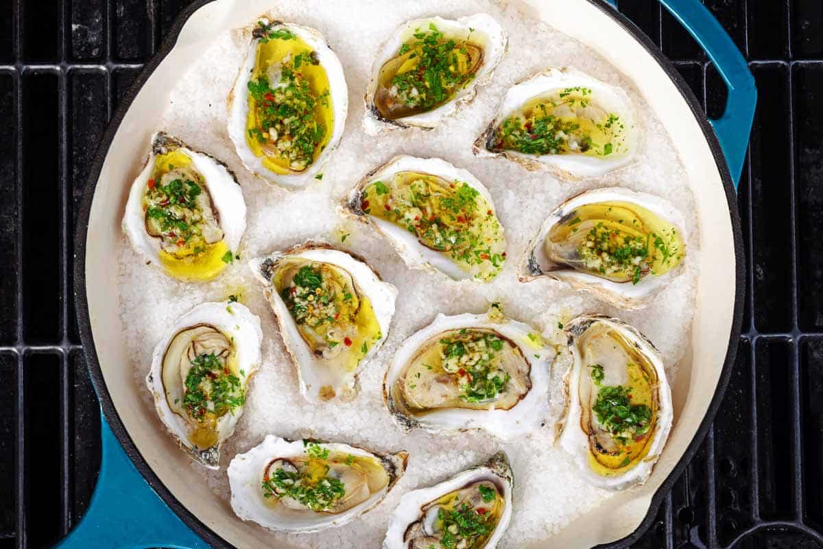 An overhead photo of several grilled oysters with garlicky herb sauce on a bed of rock salt in a skillet sitting on a grill.