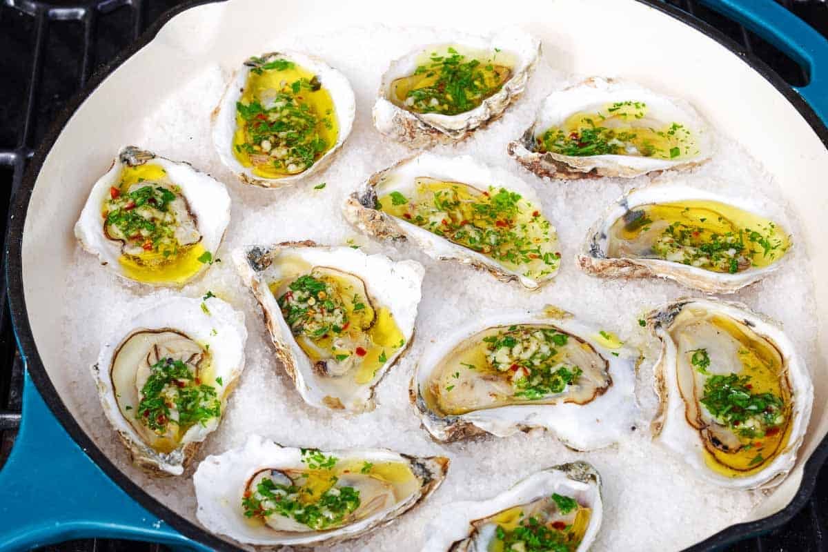 A close up of several grilled oysters with garlicky herb sauce on a bed of rock salt in a skillet sitting on a grill.