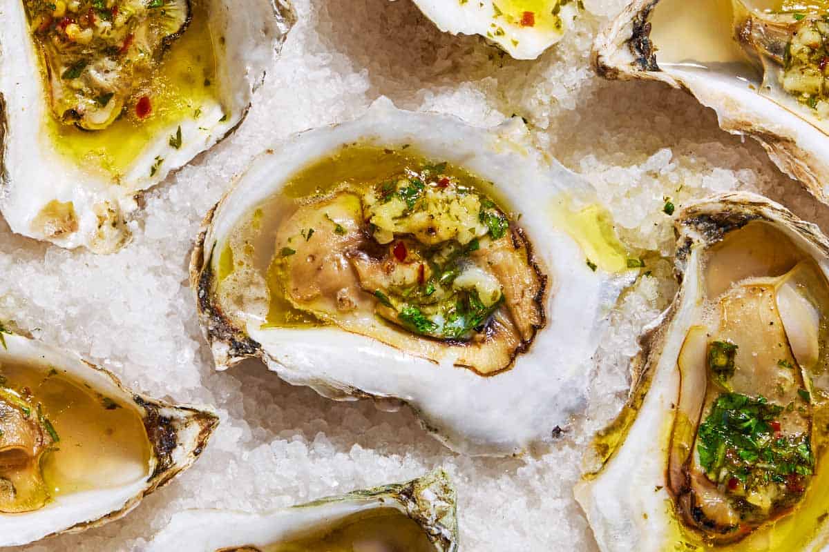 A close up of several grilled oysters with garlicky herb sauce on a bed of rock salt.