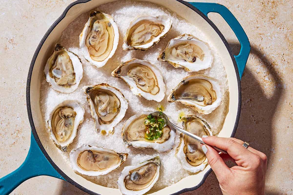 An overhead photo of the garlicky herb sauce being spooned onto shucked oysters on a bed of rock salt in a skillet.