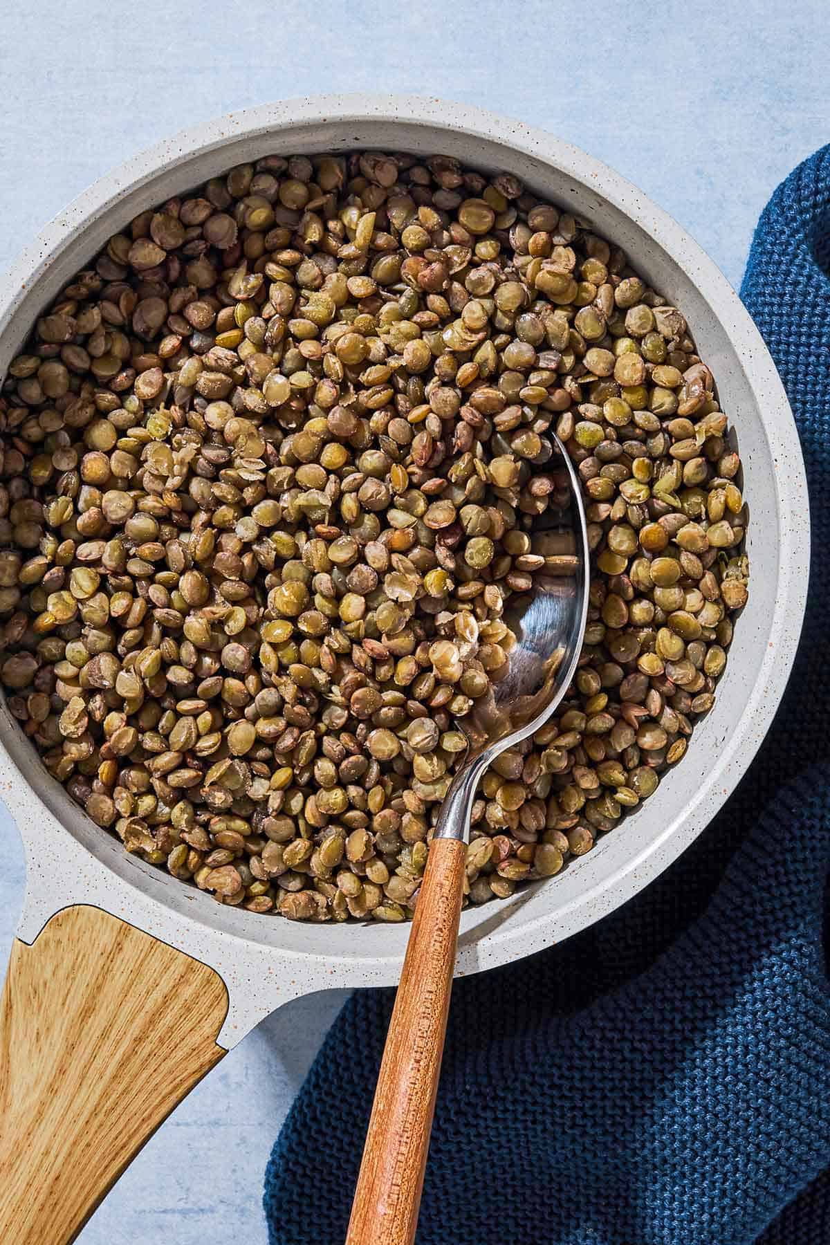 An overhead photo of green lentils in a saucepan with a spoon next to a cloth napkin.