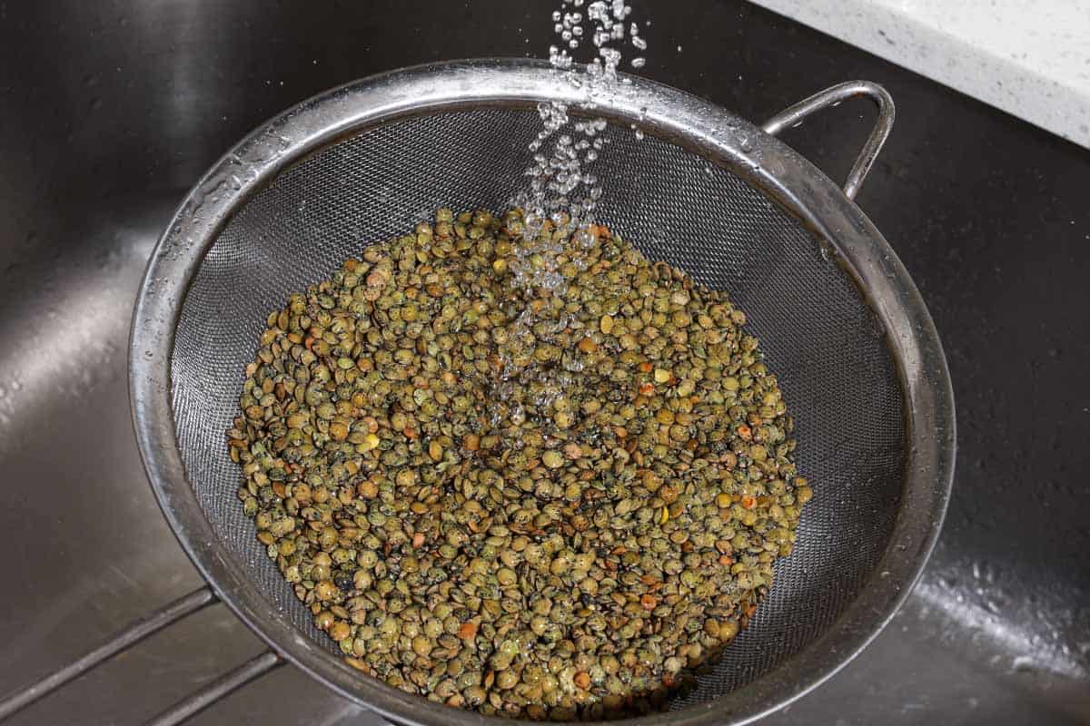 A close up of green lentils in a strainer being rinsed with water.