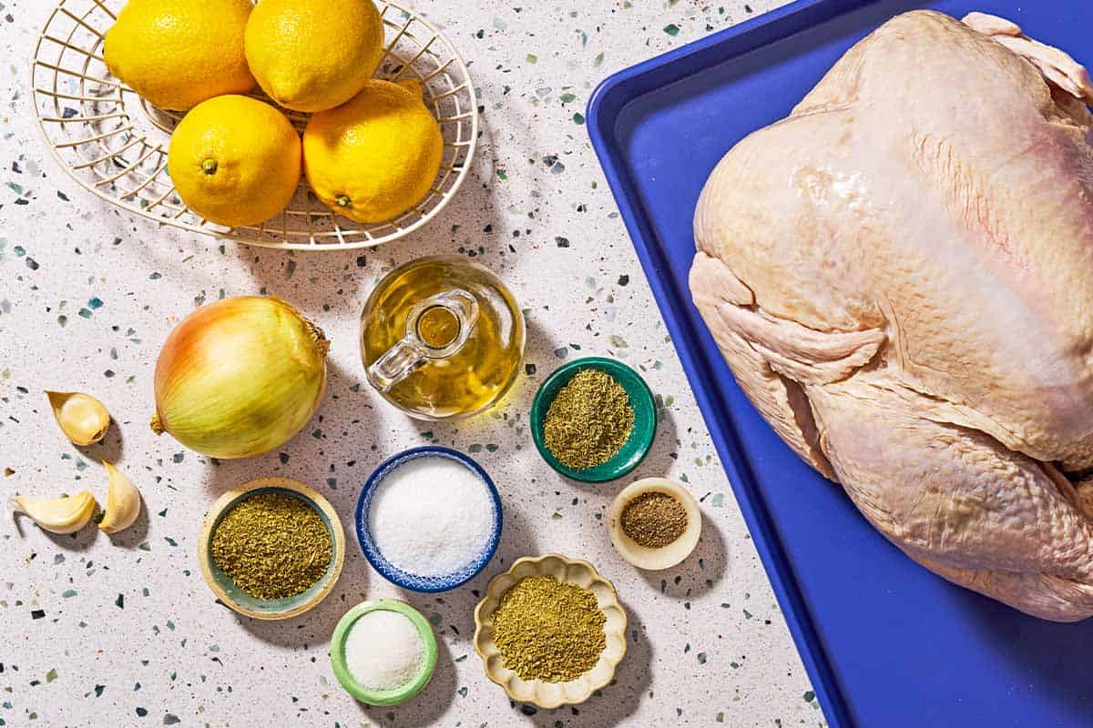 Ingredients for dry brined thanksgiving turkey including a whole turkey, garlic, olive oil, lemons, salt, oregano, fennel, thyme, sugar, black pepper, and a yellow onion.