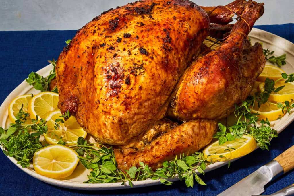 A close up of a whole roasted dry brined thanksgiving turkey on a bed of lemon wheels and greens on a platter. Next to this is a carving knife.