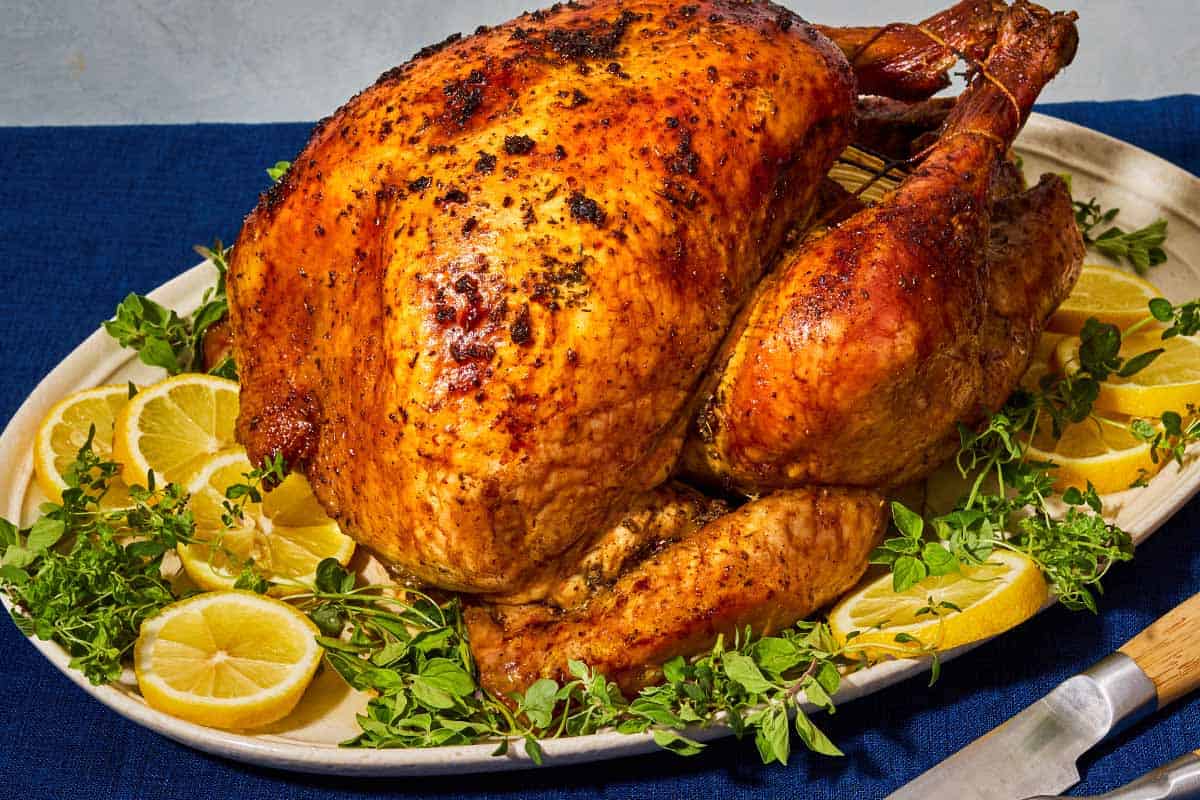 A close up of a whole roasted dry brined thanksgiving turkey on a bed of lemon wheels and greens on a platter. Next to this is a carving knife.