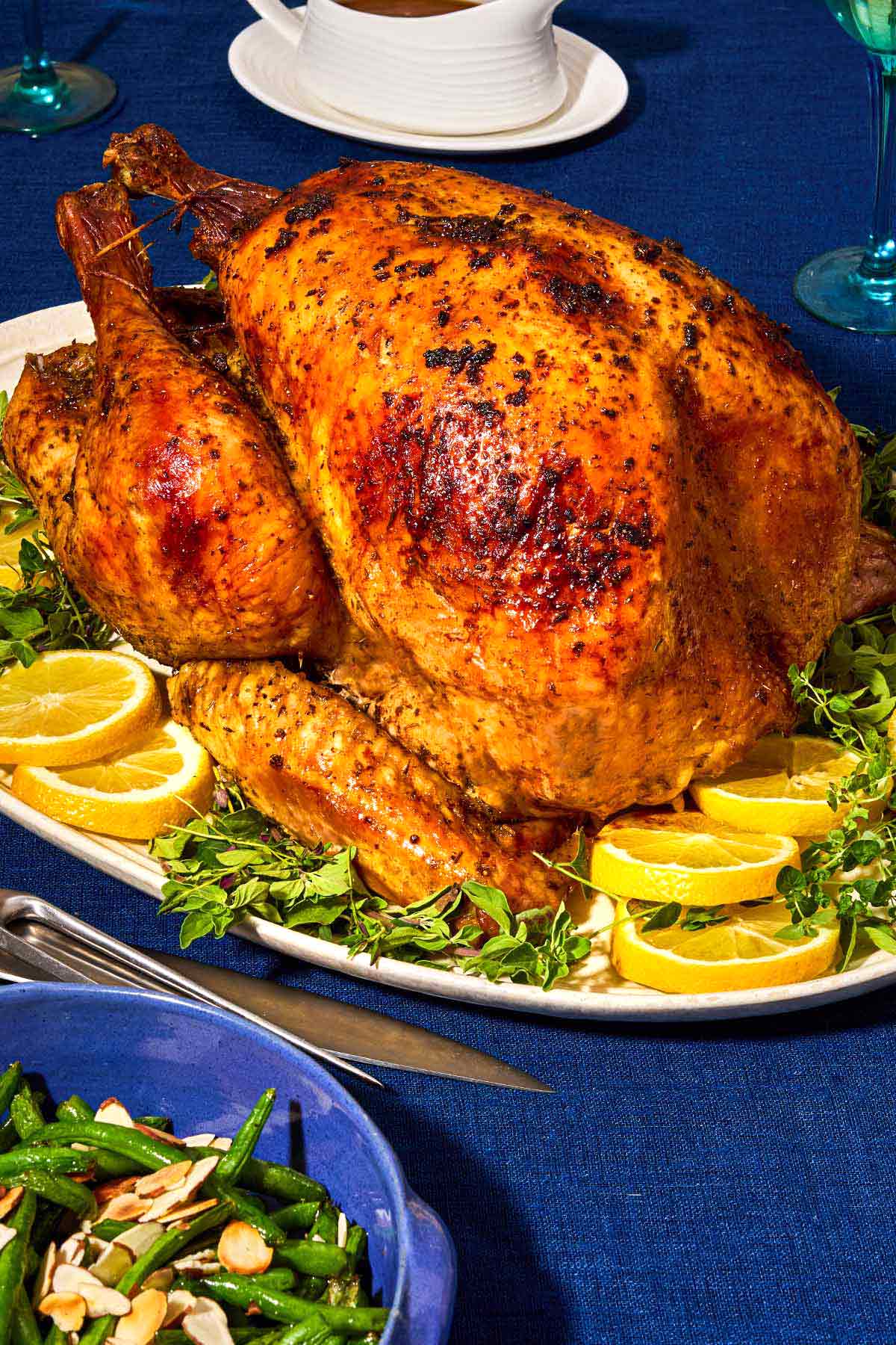 A close up of a whole roasted dry brined thanksgiving turkey on a bed of lemon wheels and greens on a platter. Next to this is a set of carving utensils, a serving bowl with green beans, and a gravy boat with gravy.