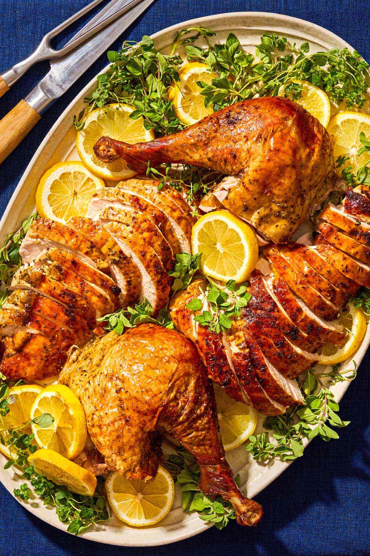 An overhead photo of sliced roasted dry brined thanksgiving turkey on a bed of lemon wheels and greens on a platter. Next to this is a set of carving utensils.