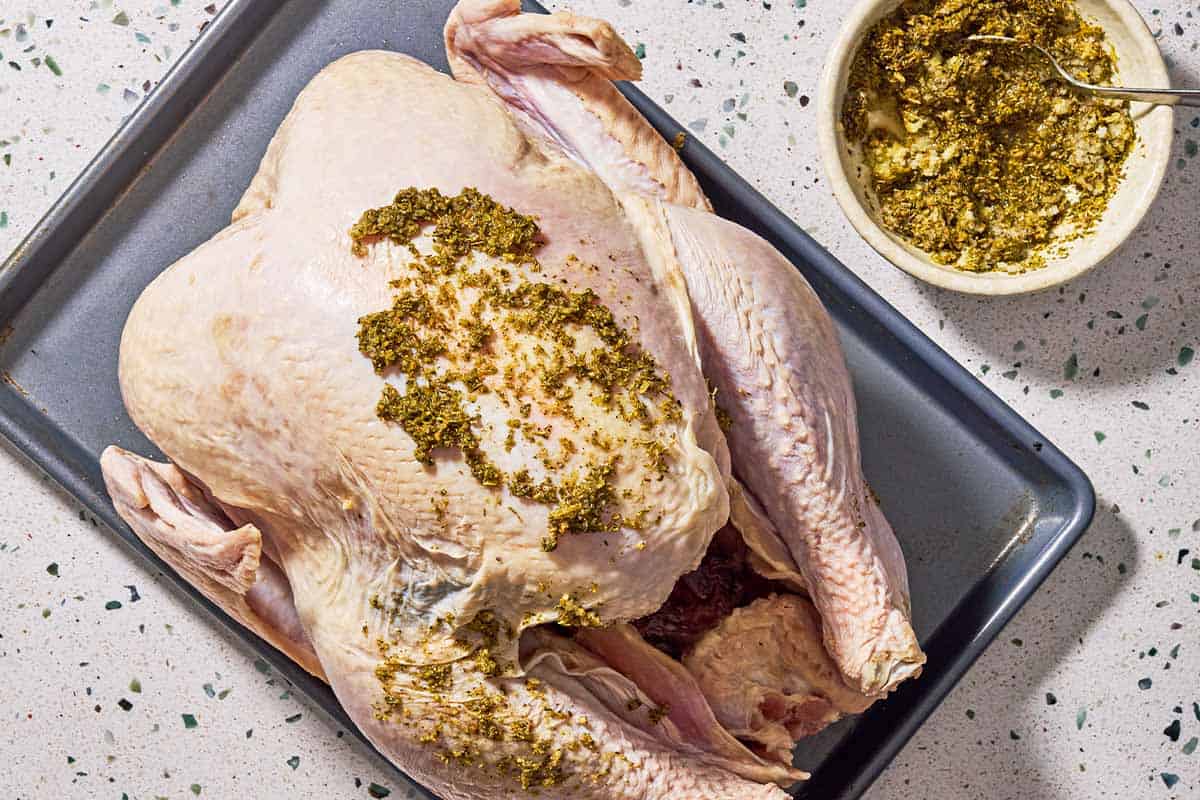 An overhead photo of a whole uncooked dry brined thanksgiving turkey being covered in the rub on a baking sheet. Next to this is a bowl of the rub in a spoon.