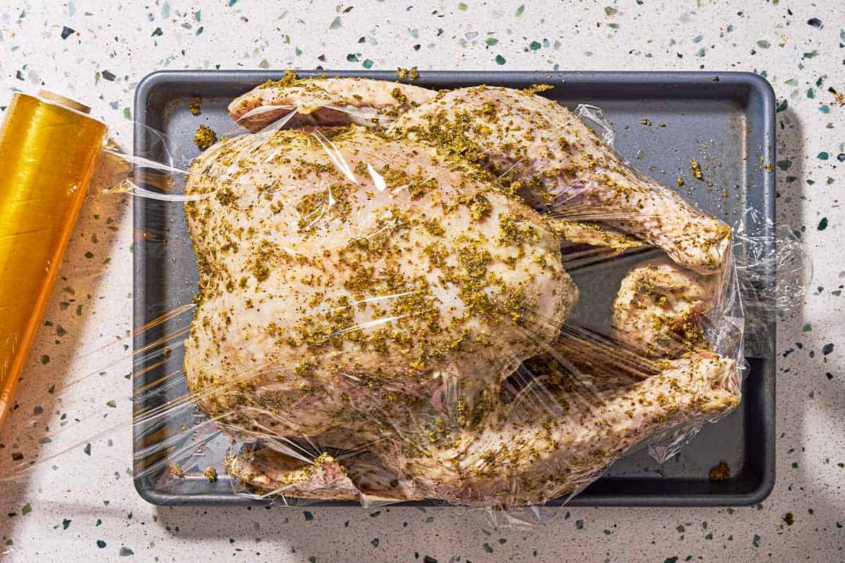 An overhead photo of a whole uncooked dry brined thanksgiving turkey covered in the rub on a baking sheet covered in plastic wrap.